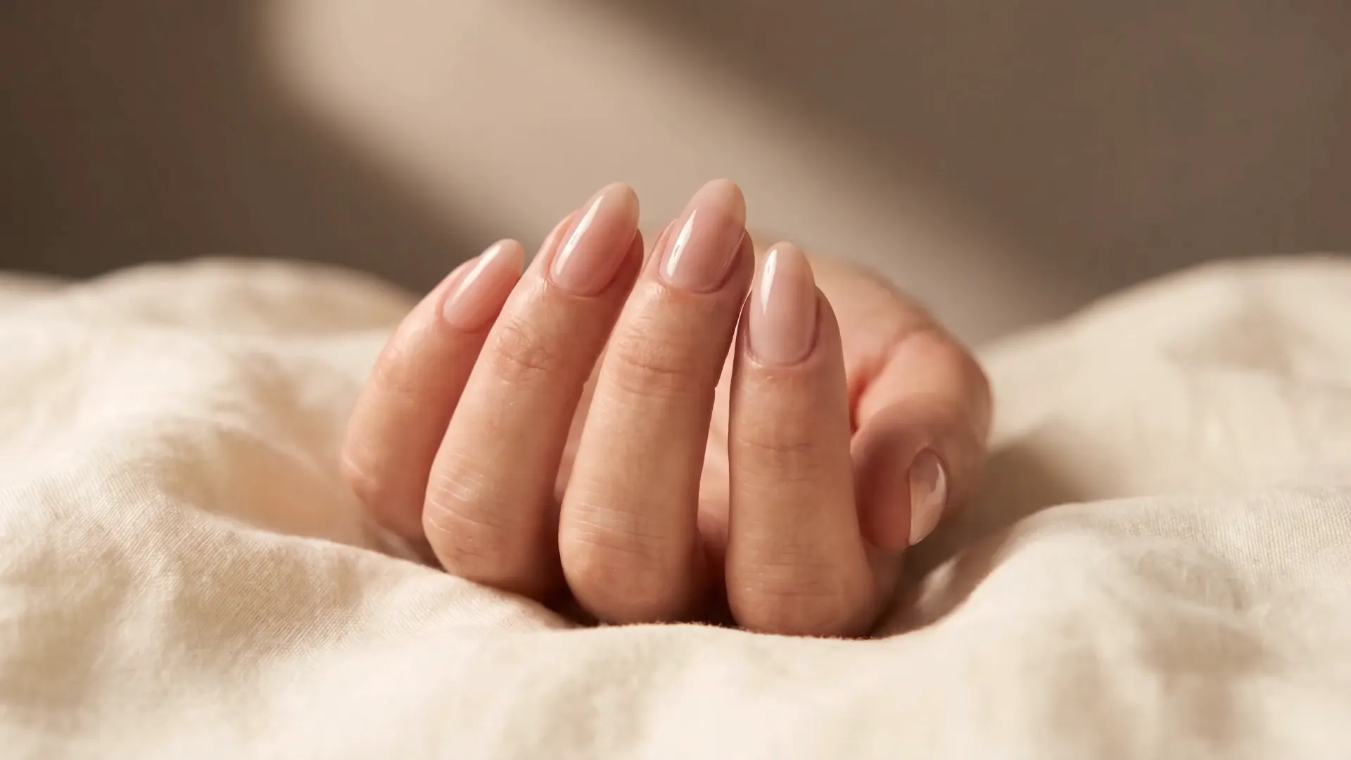 Hand with soft almond nails on cream linen as a brush applies a glossy top coat